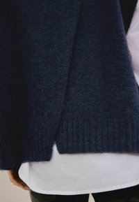 Dunkelblauer Wollpullover mit geripptem Saum, getragen über einem weißen Hemd, wobei eine teilweise Hand und schwarze Hosen darunter sichtbar sind.