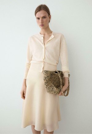 Donna che indossa un cardigan crema con bottoni e una gonna, tenendo una borsa beige texturizzata su uno sfondo chiaro uniforme.