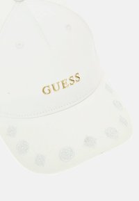 Hvid bomuldske med et guld "GUESS" logo på forsiden, med en mønstret skygge, der viser lysegrå cirkulære designs.