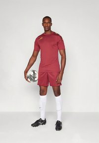 Punane sportlik särk ja lühikesed püksid, kollaste detailidega. Mängija hoiab musta-valget jalgpalli, kannab valgeid sokke ja musti krampe.