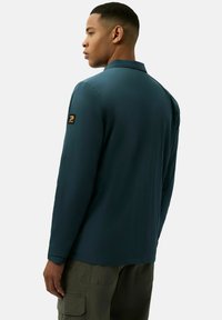 T-shirt z długim rękawem w kolorze teal z kołnierzykiem i czarną łatką logo na lewym rękawie. Gładka tekstura, zaprojektowana do codziennego noszenia.