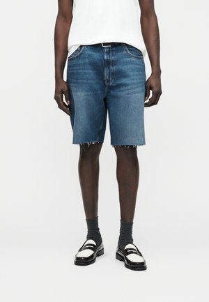 Homme portant un short en jean bleu mi-long avec ourlet brut, haut blanc, chaussettes foncées et mocassins noir et blanc, debout devant un fond blanc.