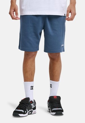 Granatowe krótkie spodenki sportowe wykonane z miękkiego materiału, z elastycznym pasem i małym logo z prawej strony na dole. Połączone z czarno-szarymi butami sportowymi.