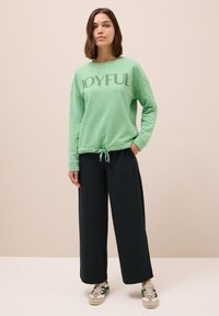 Sudadera verde menta con el texto "JOYFUL", con dobladillo ajustado, combinada con pantalones anchos oscuros y zapatillas beige-verdosas. Un atuendo simple y casual.