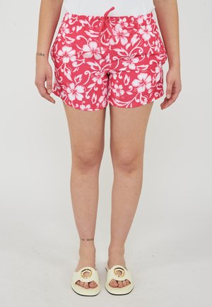 Persona che indossa pantaloncini rossi floreali con stampa di ibisco bianco, top bianco senza maniche e sandali crema aperti con dettagli dorati.