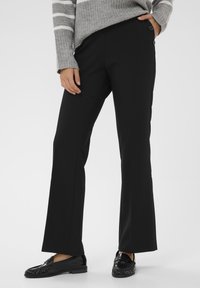 Black flared pants with a smooth texture and a fitted waist. Features two outer buttons at the waistband. Paired with glossy black loafers.