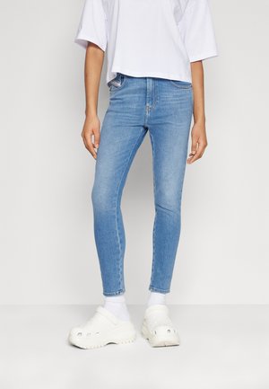 Personne portant un jean skinny bleu clair, un t-shirt blanc surdimensionné et court, des chaussettes blanches et des chaussures blanches épaisses à enfiler, debout devant un fond uni.