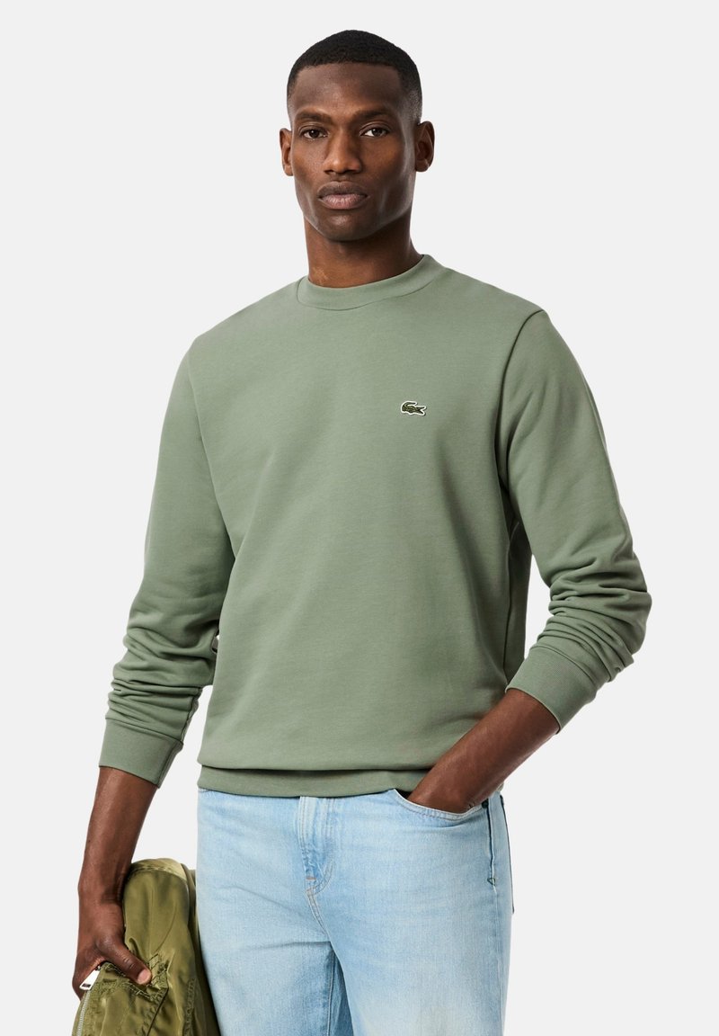 Sudadera verde de algodón con puños acanalados y cuello redondo, con un pequeño logo bordado en el pecho, combinada con vaqueros azul claro.