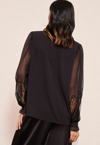 Schwarze Bluse mit transparenten Spitzenärmeln, die über ein glattes Oberteil und geraffte Manschetten verfügt. Der Rücken hat einen geraden Schnitt ohne weitere Details.
