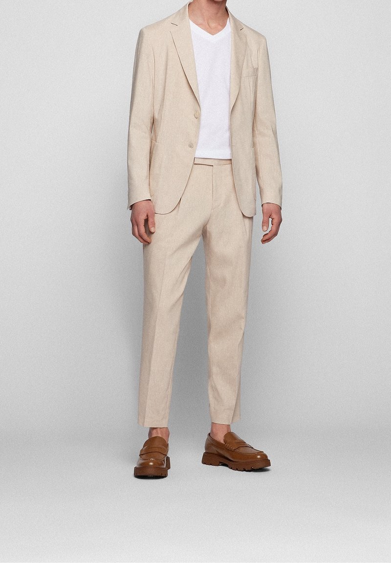 Homme portant un costume beige avec un pantalon sur mesure, une chemise blanche à col en V, et des mocassins en cuir marron, debout devant un fond neutre.