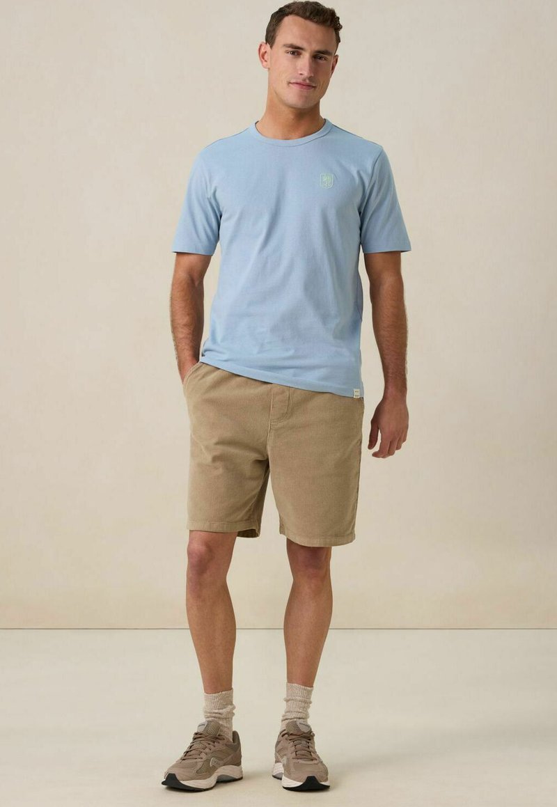 Lichtblauwe katoenen t-shirt met korte mouwen en een ronde halslijn, gecombineerd met beige shorts. De outfit bevat neutrale sokken en tan sneakers.