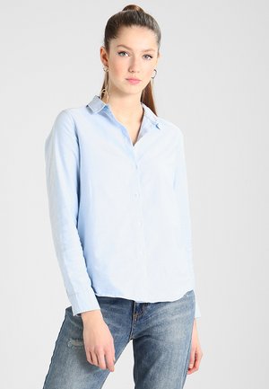 Chemise boutonnée à manches longues de couleur bleu clair avec un col et une coupe décontractée, assortie à un jean bleu délavé. Texture de tissu lisse.