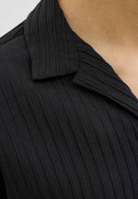Haut noir à col avec un tissu texturé présentant des rayures verticales, affichant une finition lisse et un éclat subtil.