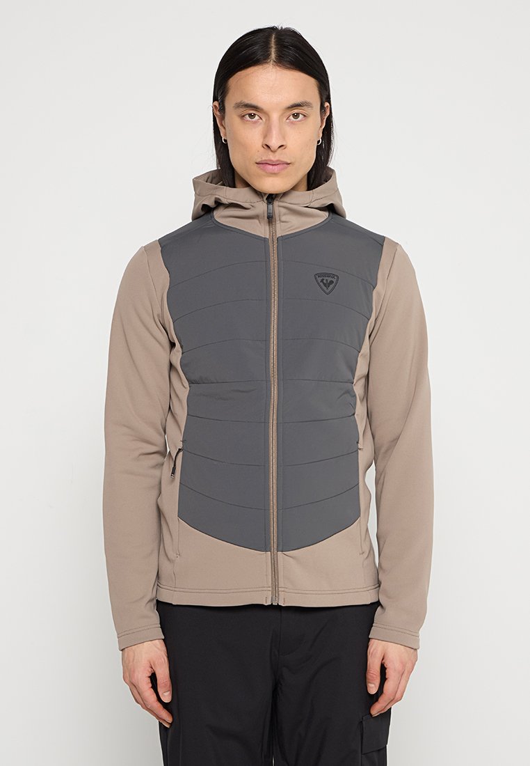 Kurtka z kapturem w odcieniach beżu i szarości, z ocieplaną przednią częścią, gładkim materiałem, pełnym zapięciem na zamek oraz akcentem logo na klatce piersiowej.