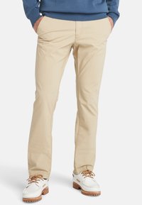 Chino di cotone beige con taglio dritto, tasche frontali e dettagli di cucitura a vista, abbinati a scarpe bianche con accenti marroni.