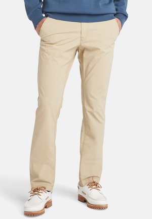 Chino di cotone beige con taglio dritto, tasche frontali e dettagli di cucitura a vista, abbinati a scarpe bianche con accenti marroni.
