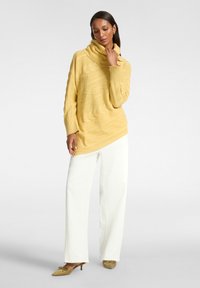 Maglione giallo oversize a maniche lunghe con collo a cascata, caratterizzato da un motivo a trecce testurizzato, abbinato a pantaloni bianchi a gamba larga e tacchi verdi.