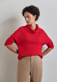 Maglione rosso a coste con collo a cowl e maniche arrotolate, abbinato a pantaloni beige. Texture liscia, vestibilità rilassata.