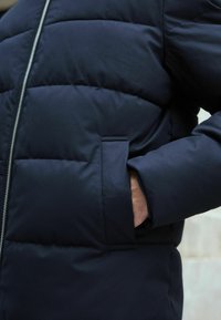 Jachetă căptușită de culoare albastră marină cu o textură netedă, având un fermoar frontal și detalii ale buzunarelor laterale. Mâneca are un design puternic și ajustat.