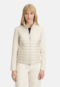 Beżowa ocieplana kurtka z poziomym pikowanym wzorem na g�órnej części, z zamkiem błyskawicznym oraz miękkimi rękawami, połączona z jasnymi spodniami.