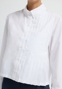 Camicia bianca con colletto, dotata di chiusura a bottoni, caratterizzata da pince verticali su un lato e una tasca a toppa, realizzata in tessuto liscio.
