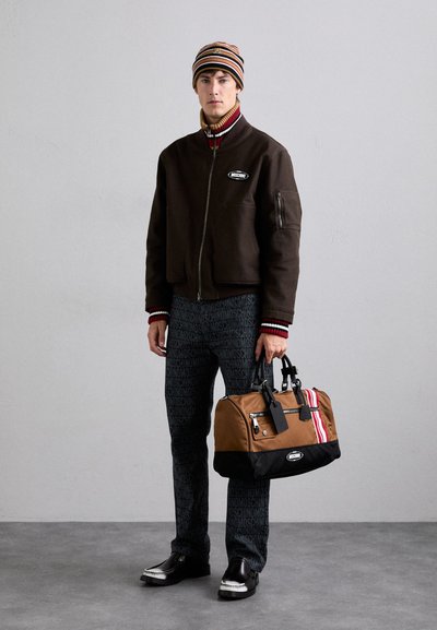 Brown bomber jacket with red and white striped cuffs, black patterned pants, and a tan duffel bag with red and white accents. Black shoes.