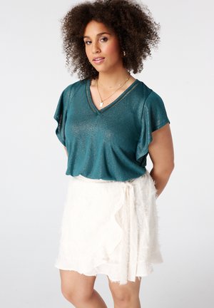 Femme aux cheveux bouclés portant un haut turquoise à manches courtes et une jupe blanche texturée, debout devant un fond clair uni.