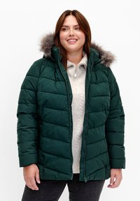 Giacca imbottita verde con cappuccio foderato in pelliccia, dotata di chiusura con zip e un maglione in maglia testurizzata sotto. Indossata con pantaloni scuri.