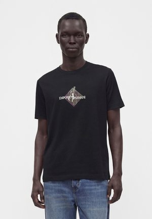 Muž v čiernom tričku Emporio Armani s maskáčovým diamantovým logom a modrých džínsoch stojaci pred jednoliatym svetlým pozadím.