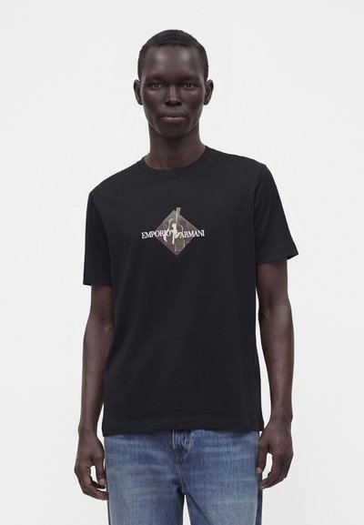 Άνδρας που φοράει μαύρο t-shirt Emporio Armani με λογότυπο ρόμβου καμουφλάζ και μπλε τζιν, στέκεται μπροστά σε απλό φωτεινό φόντο.