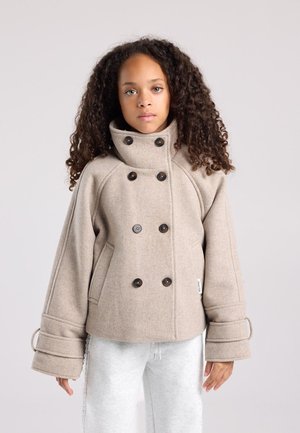 Jeune personne aux cheveux bouclés portant un manteau beige croisé à larges manches et un pantalon de survêtement blanc, debout devant un fond uni.