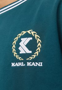 T-shirt in cotone color teal con logo ricamato bianco e oro, accompagnato da una corona d'alloro e il testo "KARL KANI" sopra l'emblema.