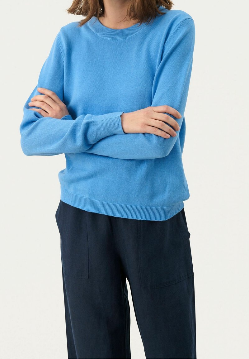 Personne portant un pull bleu clair à manches longues avec des poignets côtelés et un pantalon ample foncé, les bras croisés au niveau de la poitrine.
