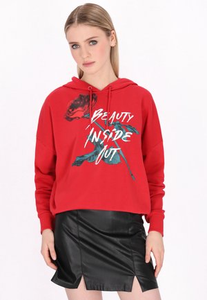 Junge Frau trägt einen roten Hoodie mit einer Rose und dem Text „Beauty Inside Out“, kombiniert mit einem schwarzen Lederrock mit vorderen Schlitzen.