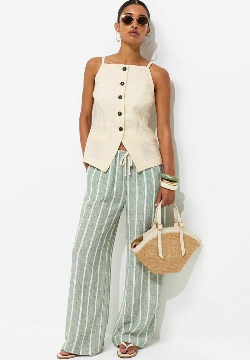 Woman wearing beige sleeveless buttoned top, green and white striped wide-leg pants, sunglasses, bracelets, holding a straw handbag.