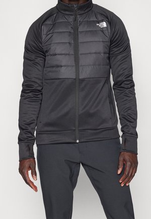 Mężczyzna noszący czarną kurtkę North Face na zamek z pikowanym panelem na piersi i otworami na kciuki w rękawach, w połączeniu z czarnymi spodniami.
