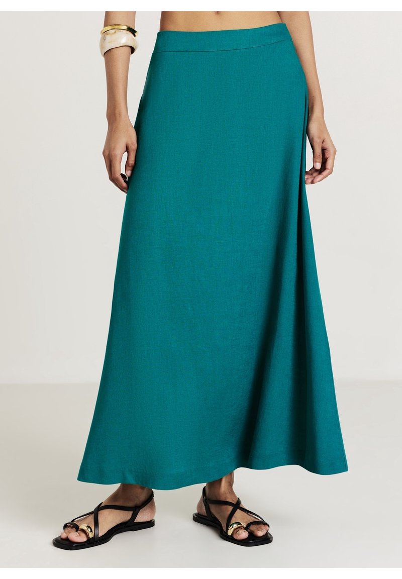 Jupe bleu sarcelle longueur cheville portée avec des sandales plates noires à brides et des bracelets dorés et blancs au bras gauche.