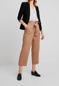 Pantalones marrones cortos con pliegues y cintura atada, combinados con una blusa floral y una chaqueta negra entallada. Unos zapatos negros sin cordones completan el conjunto.
