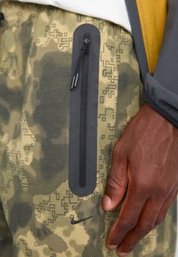 Bukser med camouflage-mønster i grønt og sort, med en slank, lynlåslomme. Har en elastisk talje og et Nike-logo.