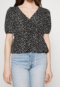 Černý peplum top s bílým tečkovaným vzorem, lehký materiál, výstřih do V a krátké nařasené rukávy. Nosený s světle modrými džíny.
