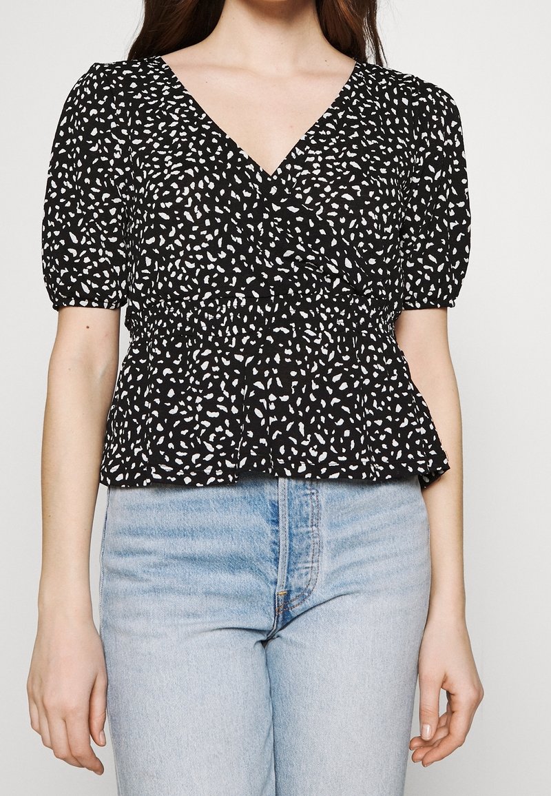 Černý peplum top s bílým tečkovaným vzorem, lehký materiál, výstřih do V a krátké nařasené rukávy. Nosený s světle modrými džíny.