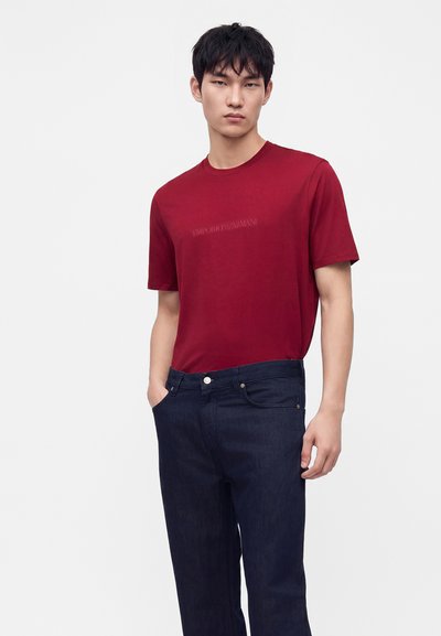 Joven con una camiseta roja de manga corta ajustada y vaqueros azul oscuro, de pie con una mano en el bolsillo frente a un fondo liso.