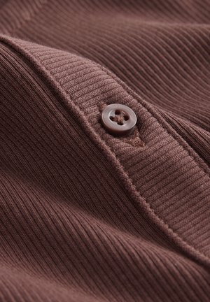 Brunt ribbat tyg med en närbild av en knappdetalj. Knappen är rund, ljusgrå med fyra hål, vilket skapar ett strukturerat utseende.