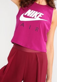 Fuchsie bezramenný crop top se velkým bílým logem Nike a textem "AIR", vyrobený z hladké bavlněné látky. V kombinaci s vysoko pasovými vínovými kalhotami.