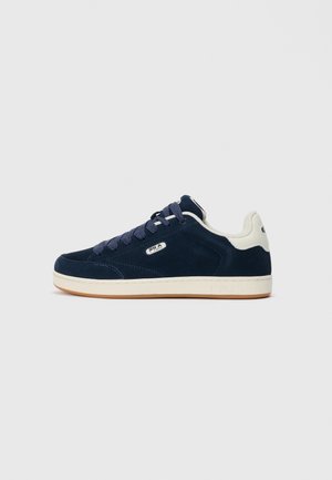 Granatowe niskie sneakersy FILA z białą łatką na pięcie, granatowymi sznurówkami i beżową podeszwą z gumową powłoką na jednolitym jasnoszarym tle.