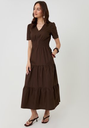 Vrouw draagt een bruine gelaagde midi-jurk met korte mouwen, V-hals en geplooide taille, staat met één hand in de zak en draagt sandalen.
