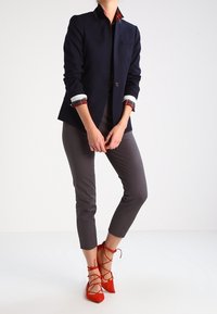 Blazer bleu marine avec col montant et fermeture à boutons, associé à un pantalon gris fuselé. Comprend des escarpins rouges à lacets et un col de chemise à carreaux.