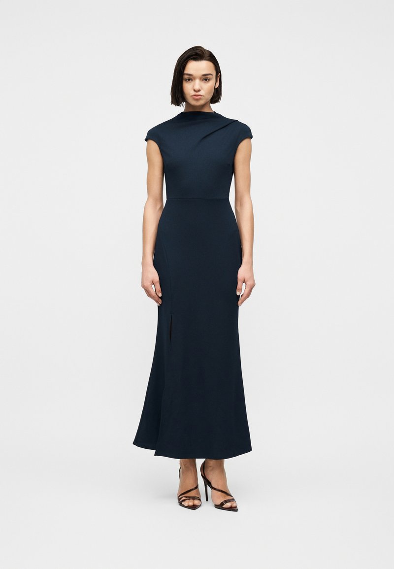 Γυναίκα με ίσια σκούρα μαλλιά που φοράει ένα αμάνικο, μακρύ φόρεμα σε σκούρο μπλε χρώμα και μαύρα πέδιλα με λουράκια, στέκεται μπροστά σε ένα απλό φόντο.