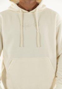 Sweat-shirt à capuche de couleur crème avec un tissu à texture en nid d'abeille, comportant une poche avant et un logo brodé discret. Sans motifs ni graphiques.