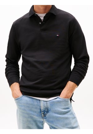 Man wearing black long-sleeve polo shirt over white undershirt, with light blue jeans, hands in pockets, against plain white background.